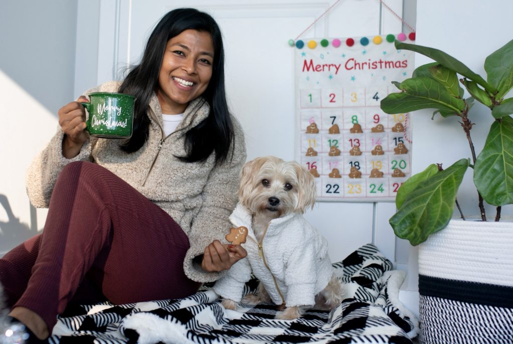 An Indian women holds a gingerbread dog cookie in front of her small tan dog. There is an advent calendar hanging in the background with gingerbread dog treats.