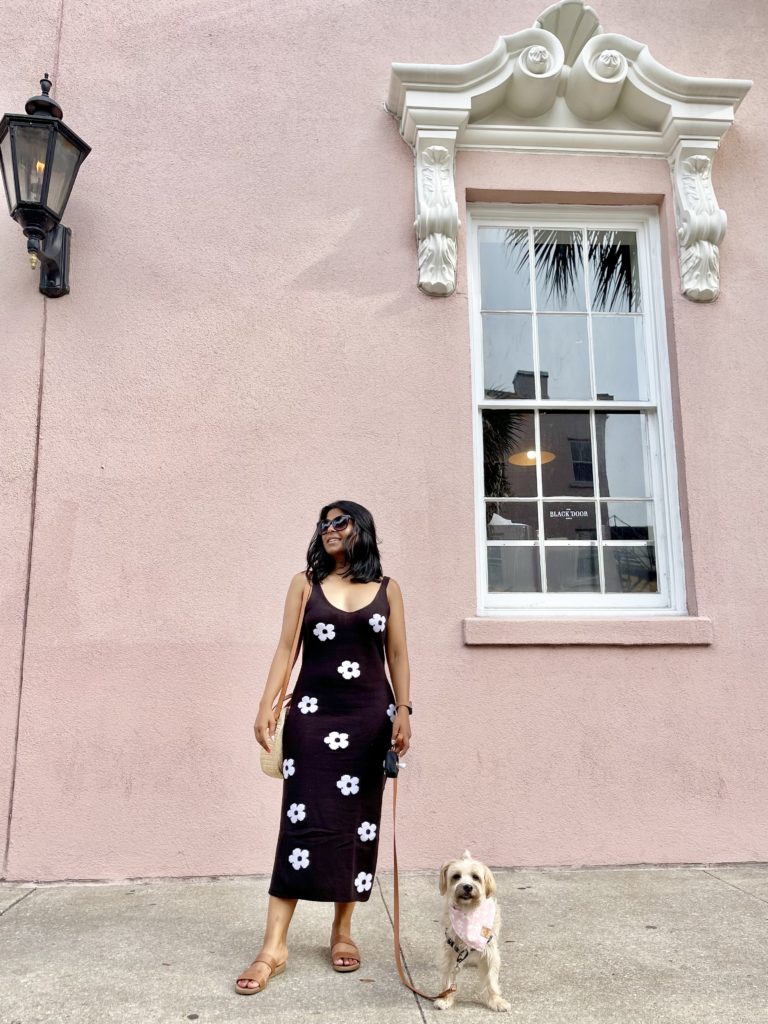 Sharon, a women of Indian descent stands with her small tan dog, Teddy in front of a pink wall in dog friendly Charleston SC
