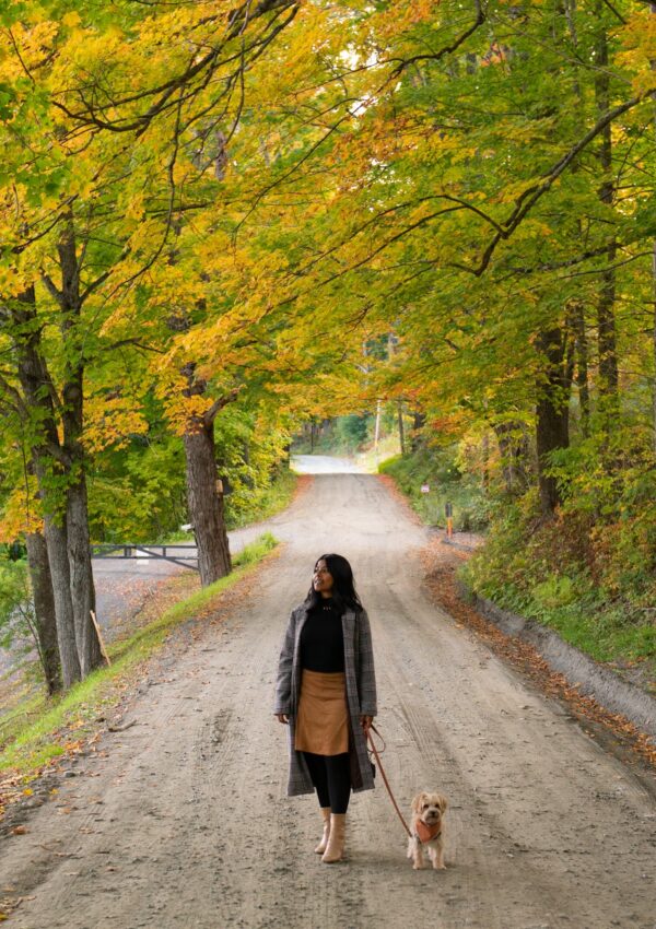sharon an asian lady stands on a dirt road with her dog Teddy in Vermont in the Fall with beautiful fall leaves on trees.